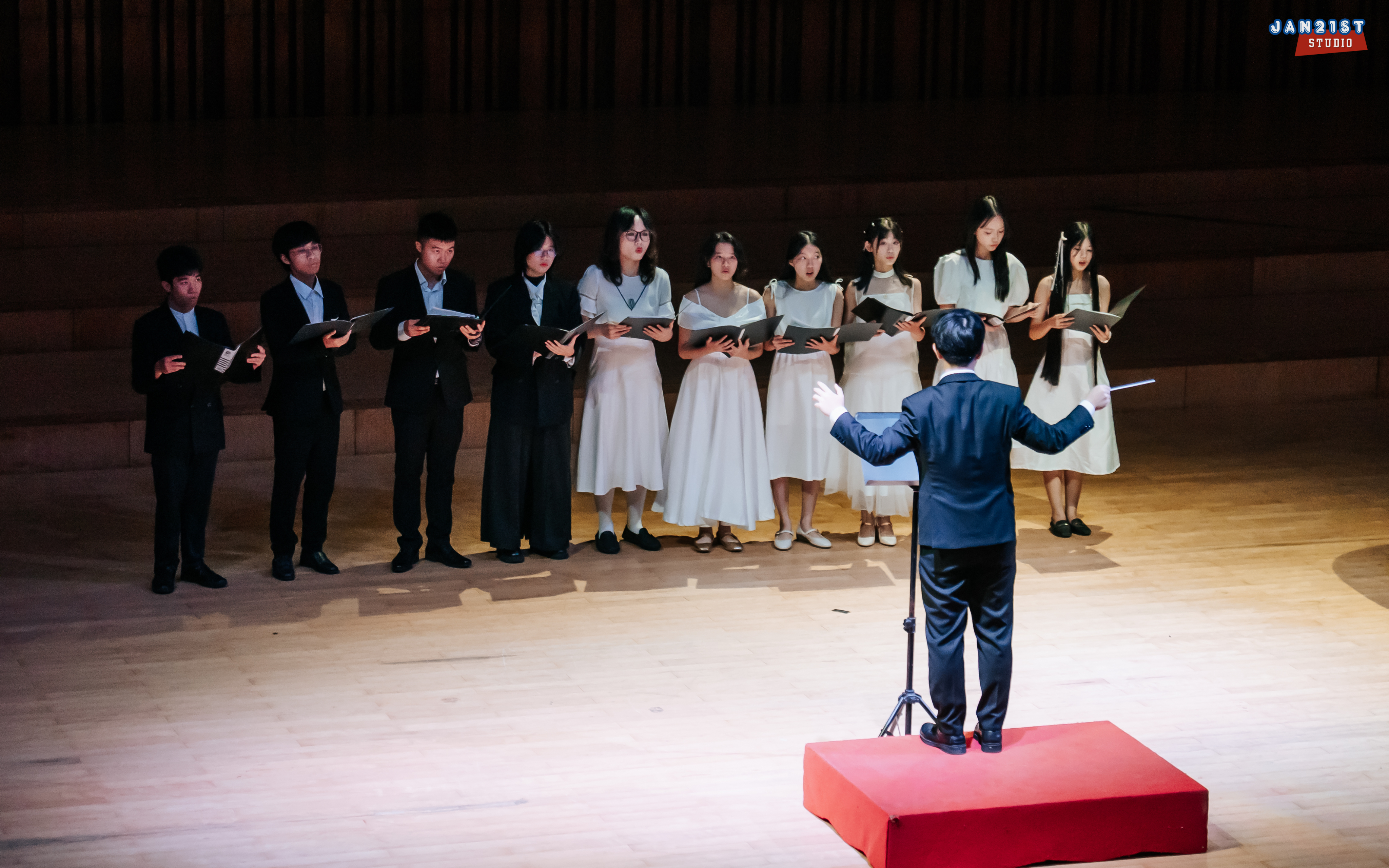 A choir in black and white facing their conductor on a wooden stage